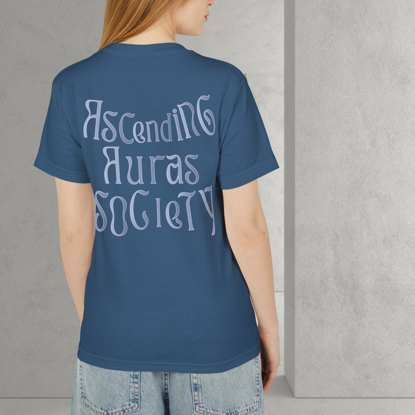 unisex t-shirt weared by american model red head clear skin the t-shirt is faded blue with the graphic saying ' ascending auras society'  she also wear a jean pants from american apparel on a grey interior background minimalist 