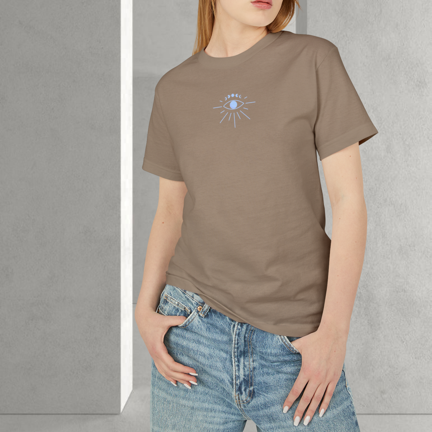 model female wearing a brown t-shirt 100% cotton with a blue silver eye on the chest printed unisex style, she stand on a interior minimalist grey background for spiritually designed brand fashion trendy 