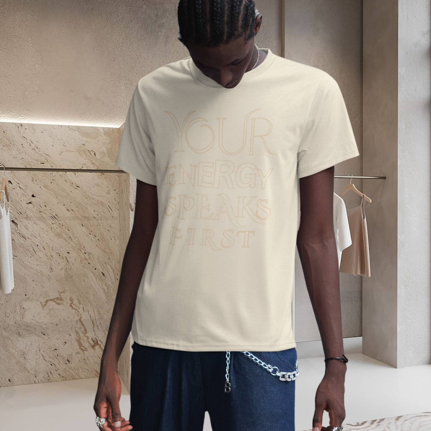 2026 trend innovative fashion concept, spiritually infused t-shirt high vibrational graphic, a african men presenting the t-shirt on the picture he is in a changing room face look down wearing the faded cream t-shirt with the elegant text graphic printed on it saying ' your energy speaks first' unisex style spiritual brand neo revelation