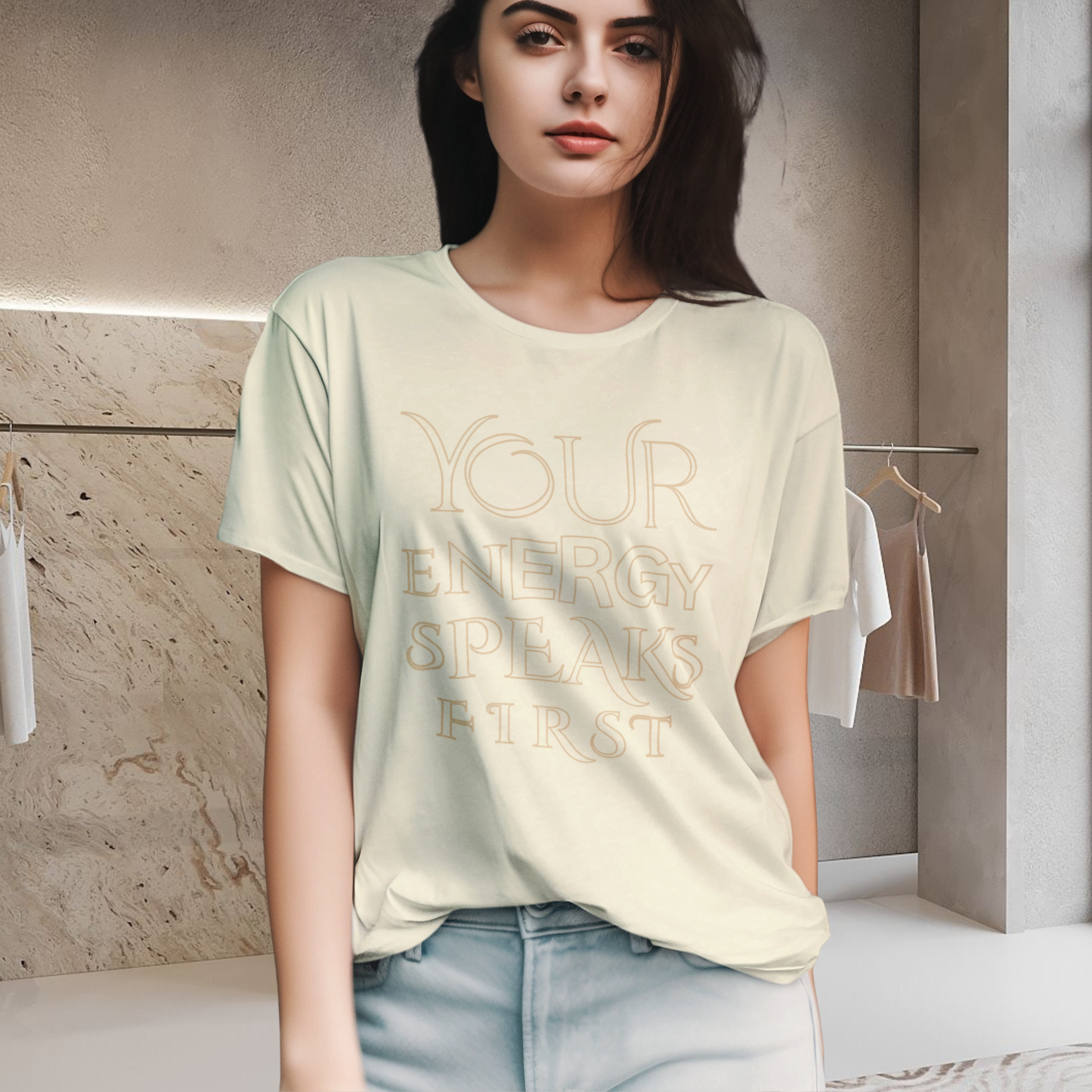 spiritually designed t-shirt high vibration graphic in elegant letters written ' your energy speaks first' in big on the front of the t-shirt, a female brunnete model wears the t-shirt with jeans pants in a changing room beige minimalist syle background neo revelation is a - spiritual innovative fashion concept -