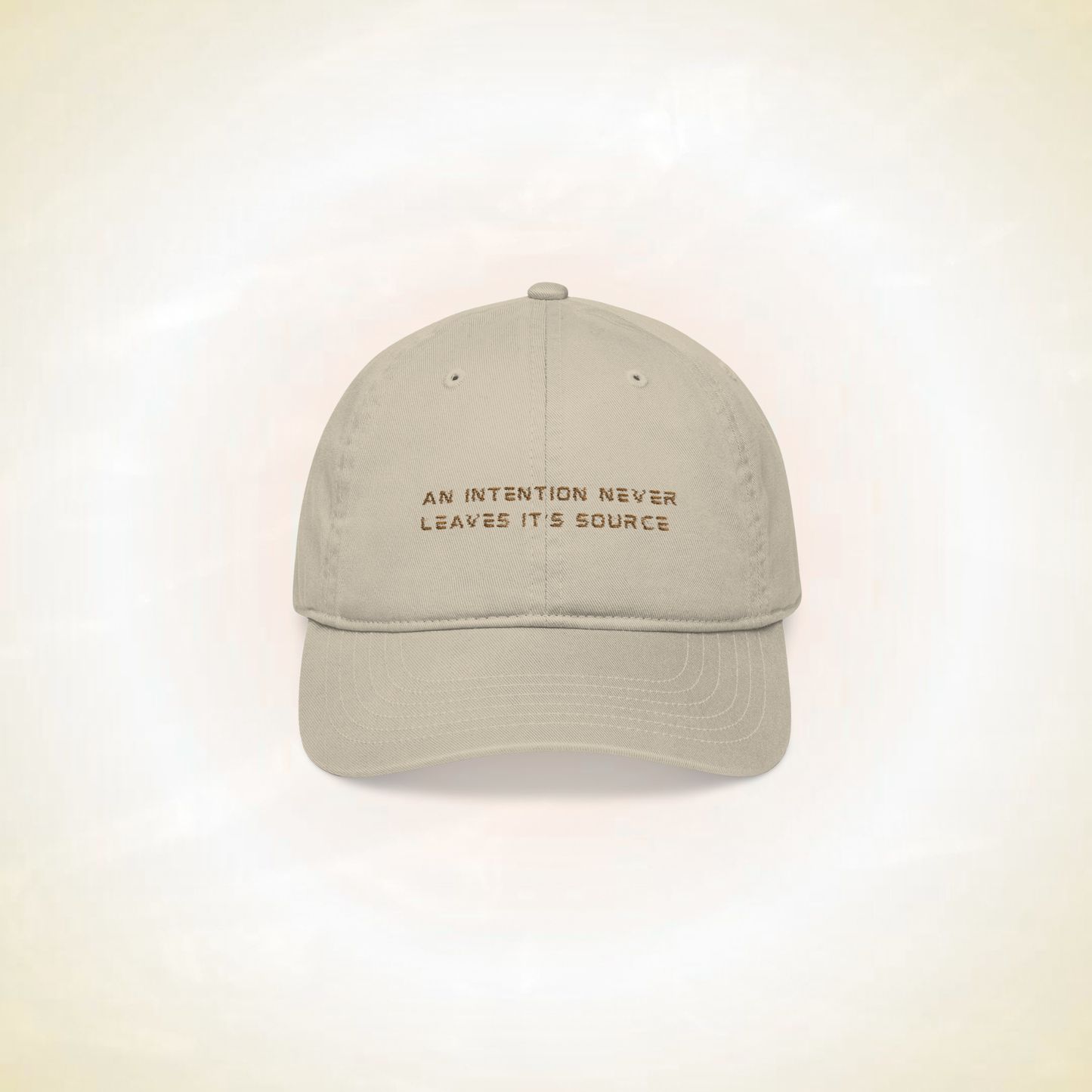 beige baseball cap with the statement ' an intention never leaves it's source' on a ligh background representing protection shield designed spiritually this hat is a crown chakra protection protecting from mental interferences, distractions, fatigue, negatives energies, innovative spiritual fashion concept for spiritual people, a solution to spiritual people problems 