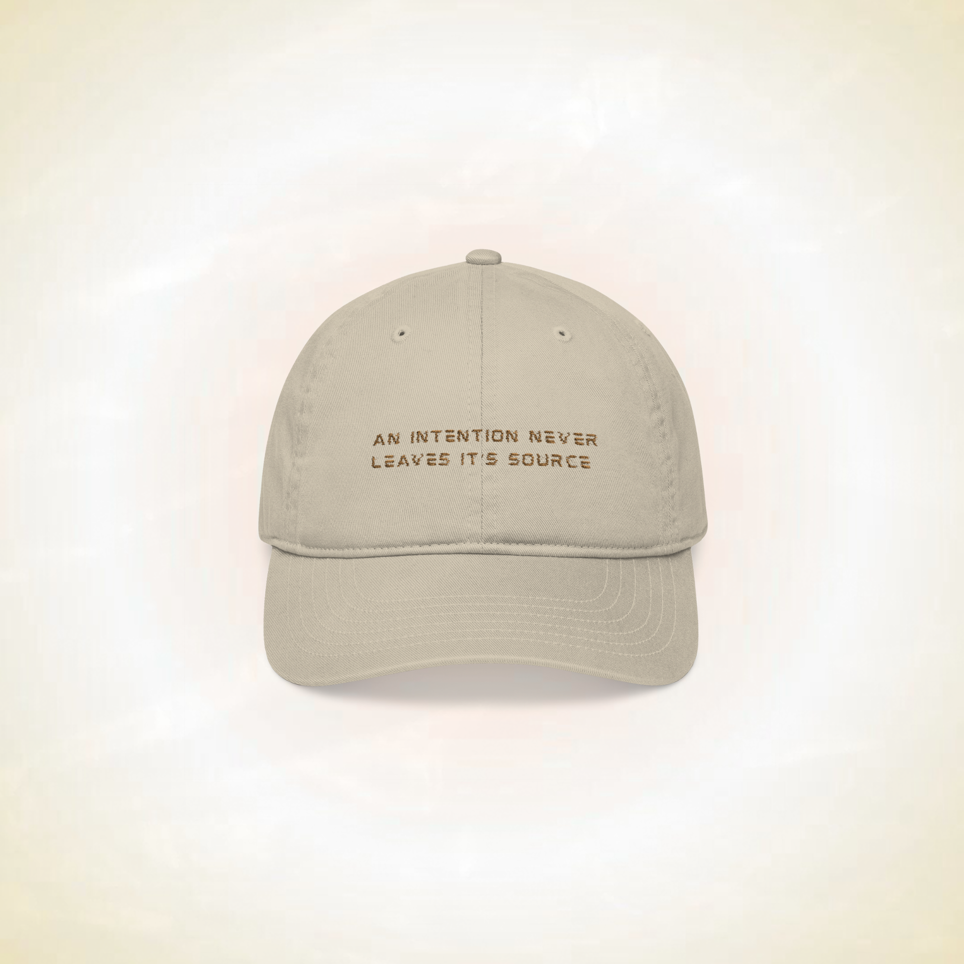 beige baseball cap with the statement ' an intention never leaves it's source' on a ligh background representing protection shield designed spiritually this hat is a crown chakra protection protecting from mental interferences, distractions, fatigue, negatives energies, innovative spiritual fashion concept for spiritual people, a solution to spiritual people problems 