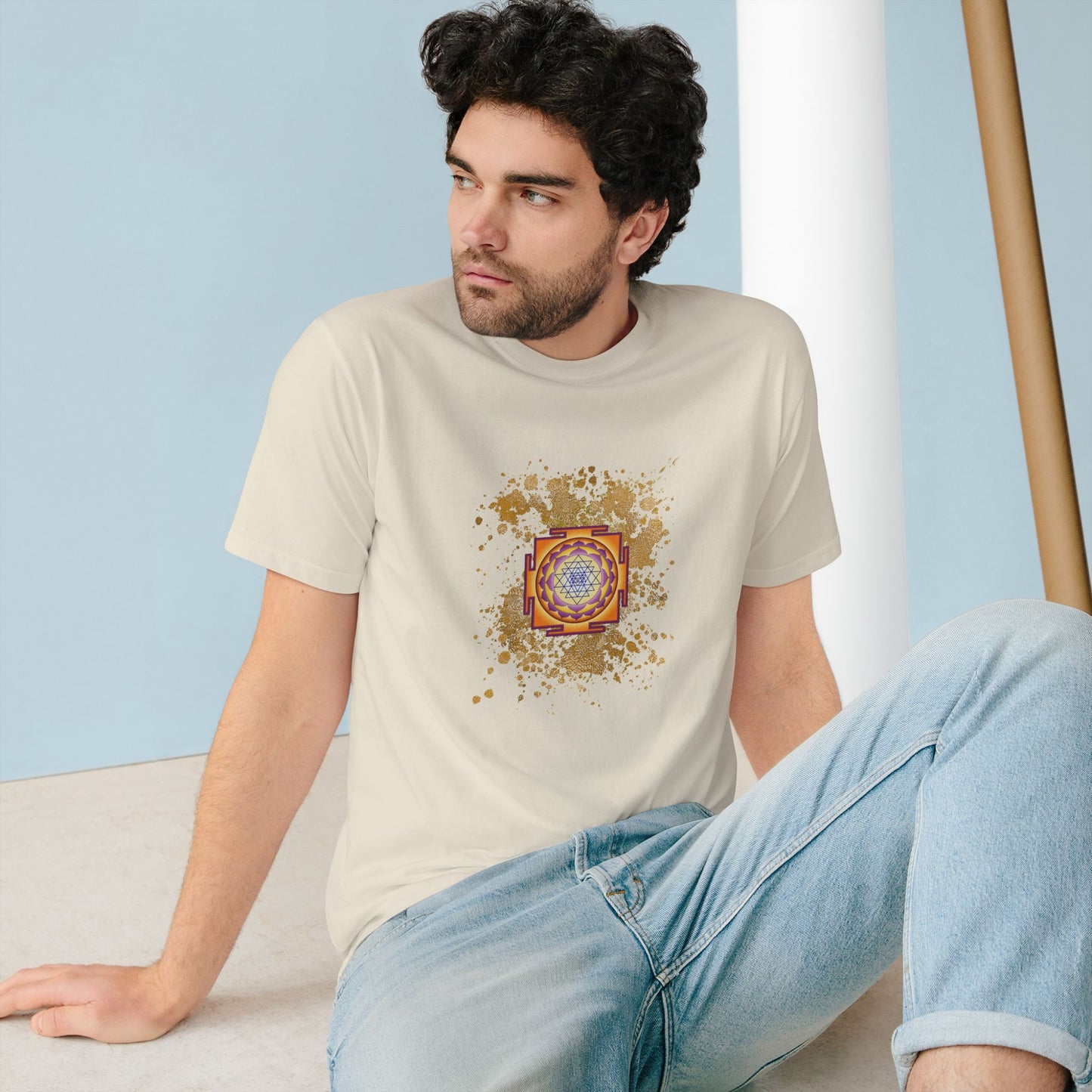 Spiritual clothing energy infused intentional fashion to wear, white cream t-shirtwith gold sparks and sri yantra graphic unisex style , the model is a european male dark hair wearing the spiritual shirt and a jean he is sate on the floor looking outside 