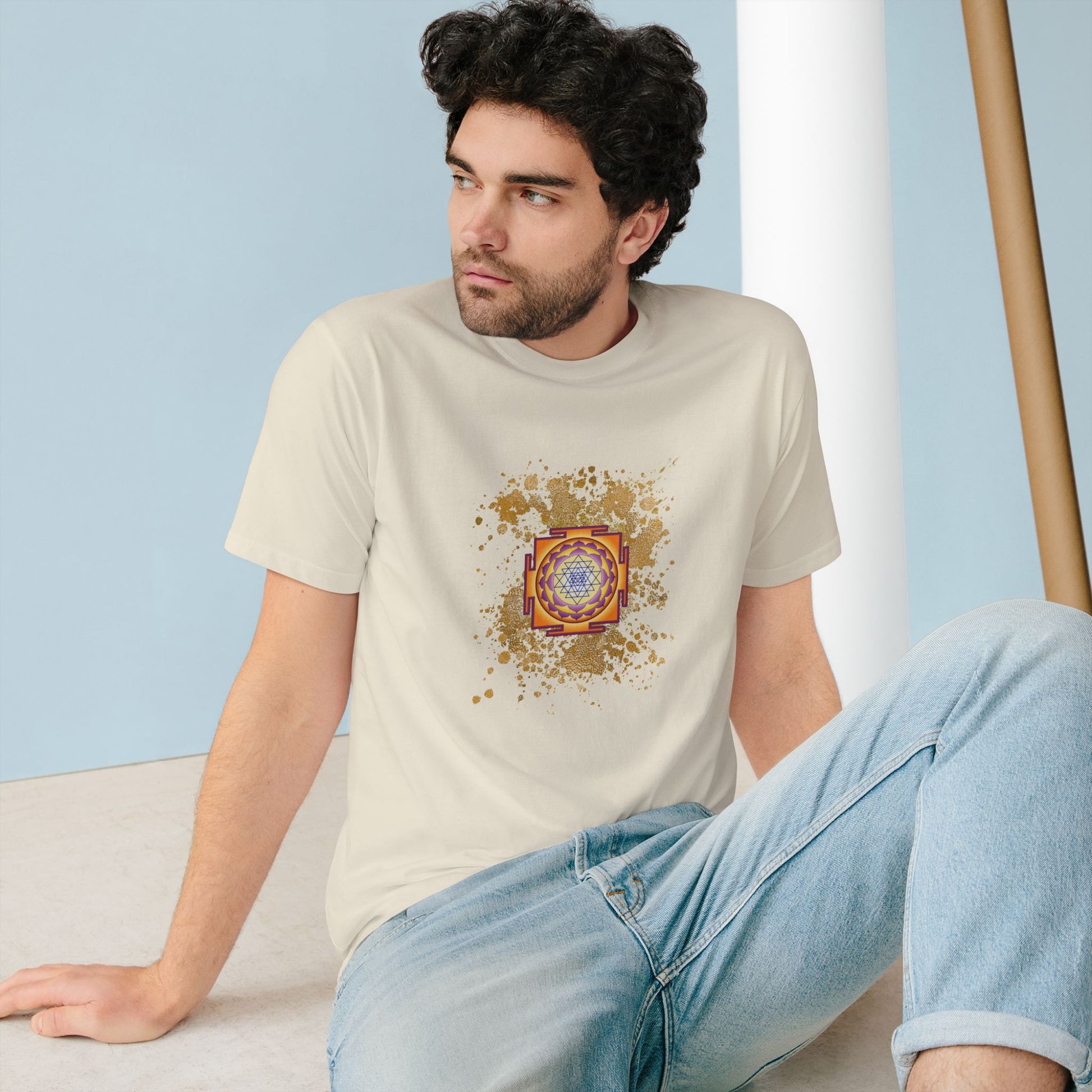 Spiritual clothing energy infused intentional fashion to wear, white cream t-shirtwith gold sparks and sri yantra graphic unisex style , the model is a european male dark hair wearing the spiritual shirt and a jean he is sate on the floor looking outside 